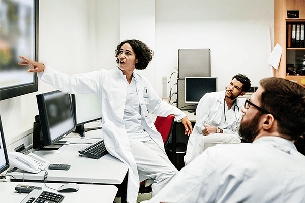 A doctor sitting on a desk, pointing to a large monitor as she has a discussion with two other doctors.