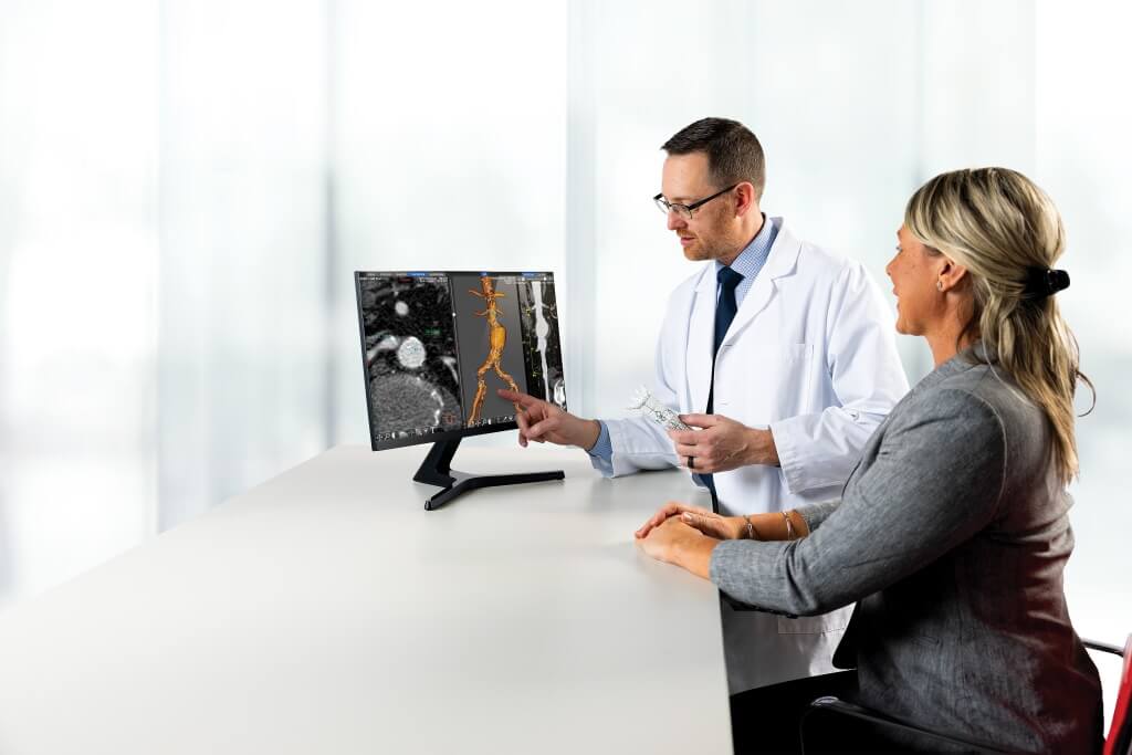 A doctor and a woman sitting at a table. The doctor is pointing at a computer screen on the table that is showing advanced imaging of the aorta.
